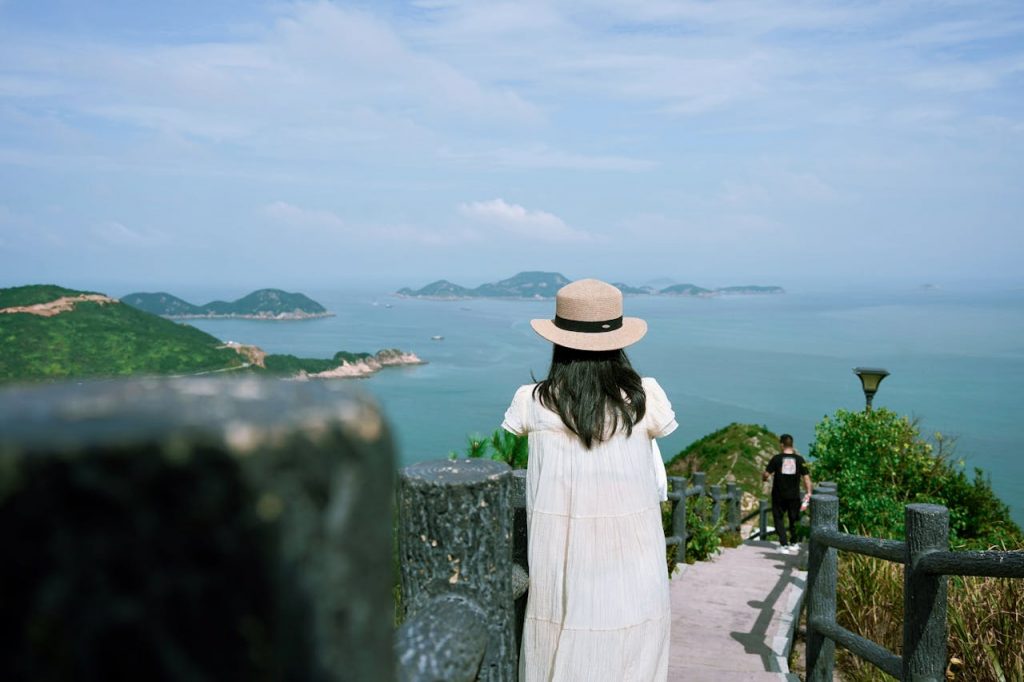 A woman in a hat gazes at the sea and islands from a hilltop path.